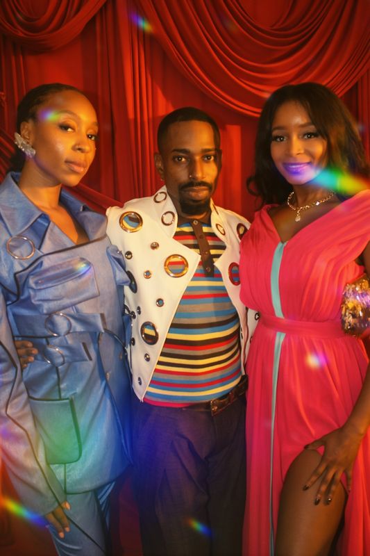 Xera Agana in a blue suit, Jet Finley in a white and gold jacket covered in mirrored circles, and Jennifer Sears in the pink archival gown, in front of a red curtain